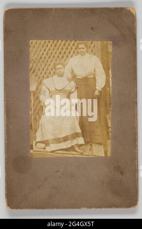 Una fotografia in bianco e nero di due donne. Una donna sta in piedi con la sua mano destra adeguata in cima alla spalla della seconda donna. La seconda donna è seduta. Le donne si trovano di fronte a un muro a rete. Una scopa è visibile a sinistra della fotografia. La fotografia è montata su carta nera. La parte posteriore del supporto è vuota. Foto Stock