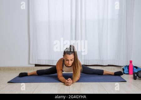 Corpo pieno di giovane donna flessibile in activewear che esegue l'esercizio laterale splits mentre allunga le gambe durante l'allenamento fitness a casa Foto Stock