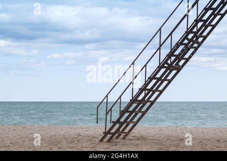 La vecchia scalinata in metallo sale sulla spiaggia sabbiosa sotto il cielo nuvoloso Foto Stock