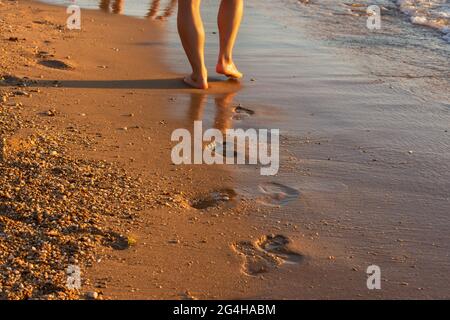 Persone spiaggia piedi da vicino. Le onde piccole si avvolgono sulla riva e sulle piste. Vacanza estiva su una spiaggia tropicale. Sfondo atmosferico al tramonto Foto Stock