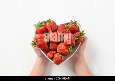 Gustoso dessert di fragola coltivata in casa biologica in mani di bambino in piatto a forma di cuore su sfondo bianco. Concept prodotti biologici. Foto Stock