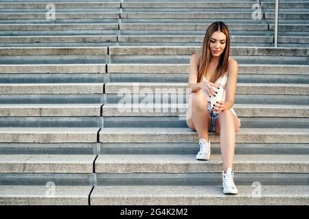 Bella ragazza, bionda con capelli lunghi, seduta sulle scale, in abiti estivi, bere caffè. Foto di alta qualità Foto Stock