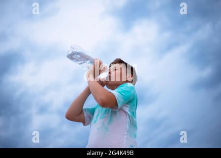 Un ragazzo contro il cielo beve acqua da una bottiglia di plastica. Sete. Calore. Cielo nuvoloso. Foto Stock