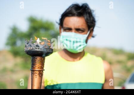 Giovane atleta con maschera medica vedendo torcia olimpica di fiamma - concetto di Olimpiadi durante la pandemia di coronavirus covid-19. Foto Stock