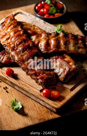 Costolette grigliate di maiale servite con pomodori ciliegini, basilico e salsa barbecue su tagliere di legno su sfondo scuro. Primo piano Foto Stock