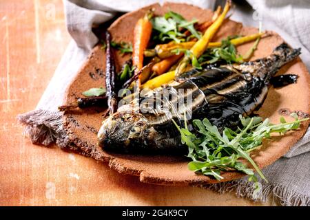 Orata fresca alla griglia o pesce dorado su piastra di ceramica avvolto in foglie di bambù servite con erbe, carote colorate, tovagliolo bianco sopra Foto Stock