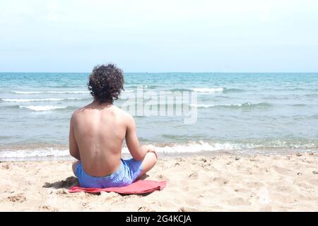 Giovane surfista con capelli ricci seduto su un asciugamano nella sabbia, con la schiena girata, guardando le onde dalla riva con un orizzonte di cielo e mare Foto Stock