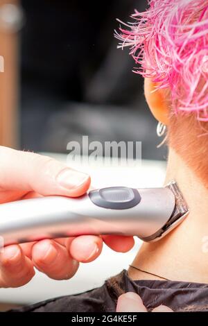 Vista posteriore della nuca e del collo per la rasatura delle mani del parrucchiere con rifinitore elettrico di giovane donna caucasica con capelli rosa corti in salone di bellezza Foto Stock