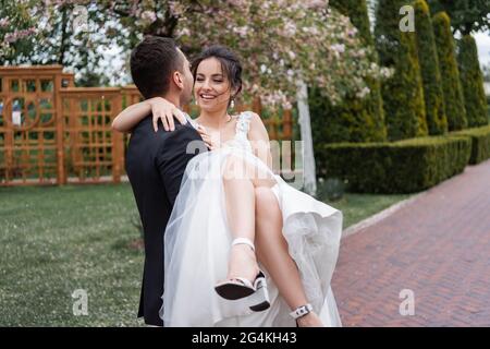 Sposo che tiene sulle mani sorridente sposa in bianco abito da sposa nel parco Foto Stock