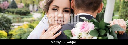 Bride allegra abbracciare lo sposo e tenere bouquet in primo piano sfocato nel parco, banner Foto Stock