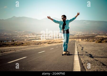 Giovane adulto che si muove su tavola lunga su una lunga strada asfaltata e all'aperto montagne sullo sfondo - concetto di libertà e persone attive Foto Stock