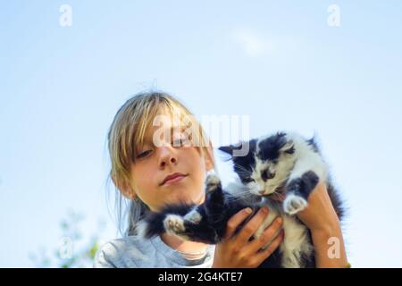 Blonde da vicino e sfocorato sorridente ragazza teen che tiene il gatto bianco e nero, gattino su sfondo blu cielo. Esterno. PET, concetto di amore animale. Fuori Foto Stock