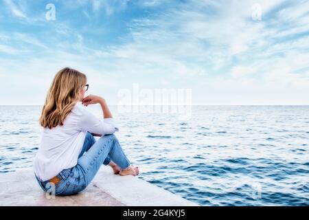 Vista posteriore di una donna che indossa abiti casual mentre si siede sul mare e si guarda al mare. Foto Stock