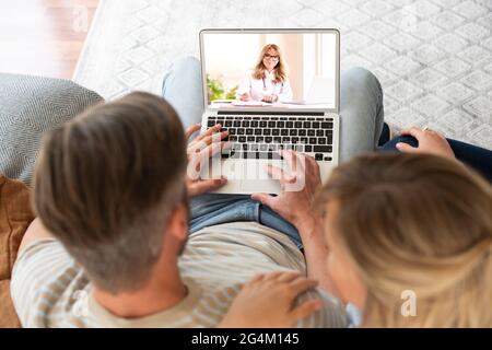 Ripresa posteriore di una coppia seduta sul divano a casa e videochiamata con il medico. Foto Stock
