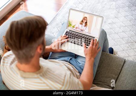 Vista posteriore di un uomo seduto sul divano a casa e videochiamata con un medico. Foto Stock