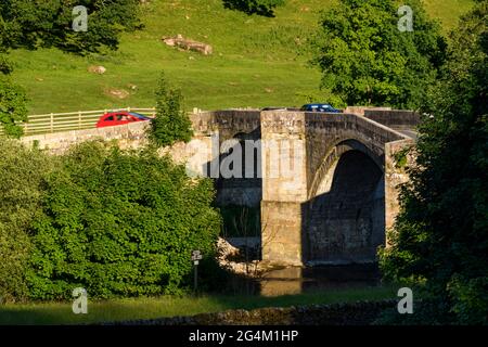 Vista panoramica sul lungofiume rurale del ponte storico, in pietra, ad arco sul fiume Wharfe (2 auto che attraversano) - Ponte di Barden, Yorkshire Dales, Inghilterra, Regno Unito. Foto Stock