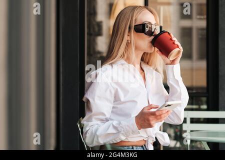 Donna elegante seduta a un tavolo da caffè e bere mentre si utilizza lo smartphone, imprenditore fidato donna indossando occhiali da sole e camicia bianca in attesa Foto Stock