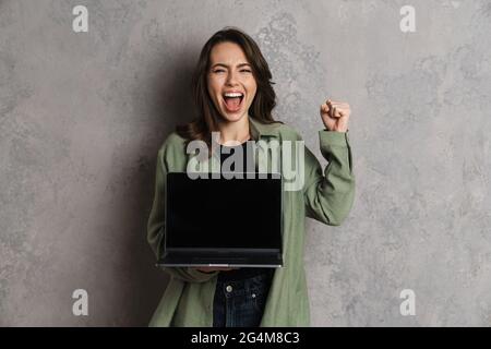 Eccitata bella ragazza bella facendo gesto vincitore e mostrando notebook isolato su parete grigia Foto Stock