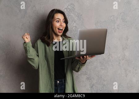 Sorpresa bella ragazza che fa gesto vincitore mentre usando il laptop isolato sopra parete grigia Foto Stock