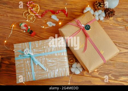 Tavolo in legno con regali di Capodanno e decorazioni natalizie. Scatole regalo in carta da imballaggio artigianale con motivi bianchi e oro. Coni, ghirlande, canne caramelle, nastro e buccia arance sullo sfondo. Foto Stock