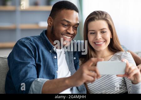 Felice coppia interracial guardare i contenuti video sullo smartphone mentre ci si rilassa a casa Foto Stock