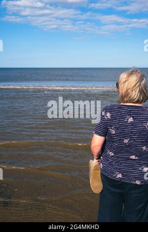 Una donna bionda ama guardare il mare mentre le onde rotolano in spiaggia in una bella giornata di sole nel Regno Unito Foto Stock