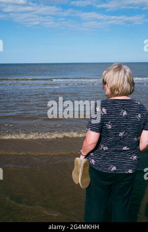 Una donna bionda ama guardare il mare mentre le onde rotolano in spiaggia in una bella giornata di sole nel Regno Unito Foto Stock