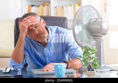 L'uomo soffre dal calore durante il lavoro in ufficio e si tenta di raffreddarsi dalla ventola Foto Stock