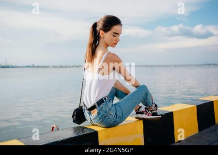 Indietro vista giovane donna con top bianco e jeans blu seduta su parapetto nero-giallo, riposante vicino al mare in giornata di sole. Foto Stock