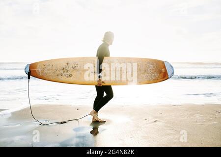 Vista laterale del surfista vestito con muta che cammina sulla spiaggia con la tavola da surf al mattino con l'alba sullo sfondo Foto Stock