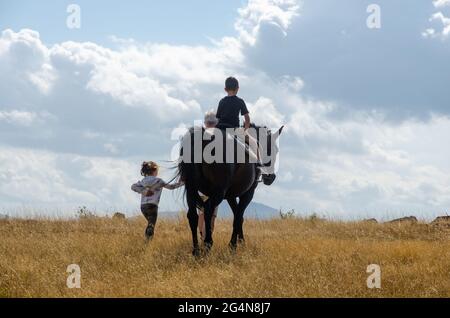 а la bambina conduce un cavallo che è guidato da un ragazzino in un campo di montagna. Foto sul retro. Foto Stock