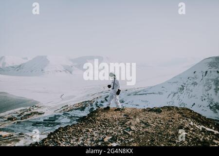 Cosmonauta irriconoscibile che indossa spacesuit bianco che si erge sul bordo di una montagna rocciosa in inverno e che ammira il paesaggio incredibile a Svalbard Foto Stock