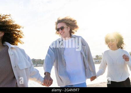 Gruppo di giovani donne e uomini diversi con capelli ricci che tengono le mani mentre si cammina sulla costa del paesaggio urbano in retro illuminato Foto Stock