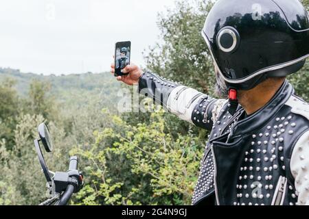 Vista laterale di un biker maschile con cuscinetti irriconoscibili in una giacca trendy in pelle con rivetti e casco protettivo che si autoritratto mentre si siede in moto Foto Stock
