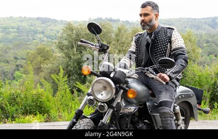 Il corpo pieno di biker maschile etnico bearded messo a fuoco in giacca di pelle nera seduta sulla moto moderna su strada asfaltata tra lussureggianti alberi verdi che crescono in Foto Stock