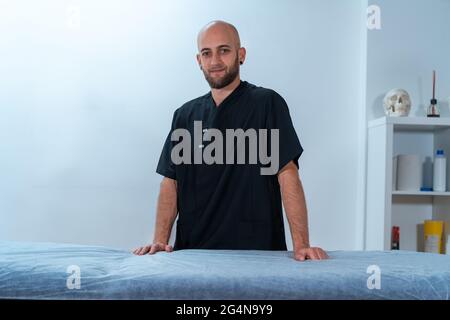 Contenuto terapeuta fisica maschile con barba guardando la fotocamera contro gli scaffali con cranio e bastoni di incenso in clinica Foto Stock