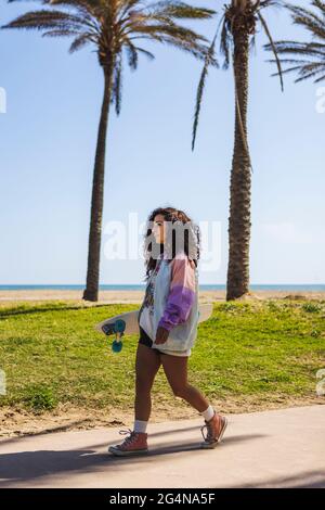 Vista laterale completa del corpo della femmina con skateboard a mano che cammina sul marciapiede lungo alte palme contro la costa e il mare Foto Stock