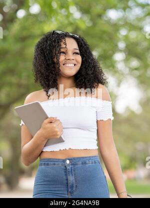Felice giovane afroamericana femmina in estate vestito a piedi tenendo tablet vicino alberi verdi nel parco di giorno Foto Stock