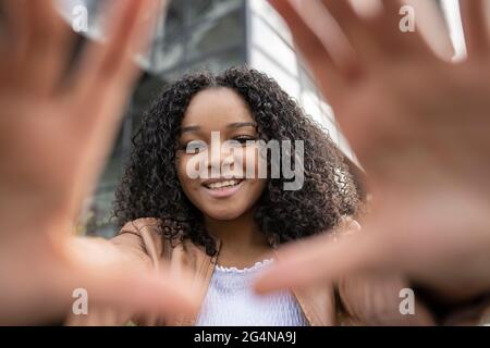 Donna afroamericana positiva con capelli ricci sorridenti e che raggiungono le mani alla macchina fotografica mentre si trova in strada vicino a un edificio moderno Foto Stock