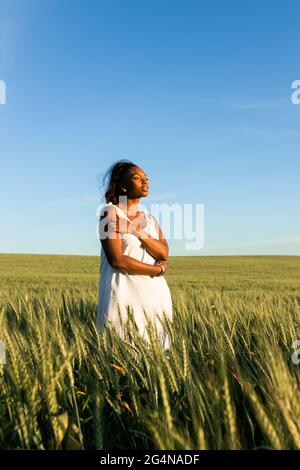 Giovane donna nera in abito estivo bianco passeggiando sul campo di grano verde mentre si guarda via di giorno sotto il cielo blu Foto Stock