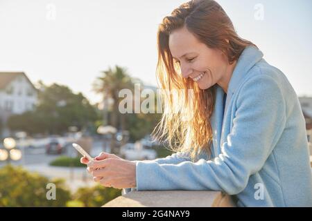 Sorridente signora adulta in caldo cappotto di navigazione telefono mentre si appoggia su recinzione vicino alla strada della città in giornata di sole Foto Stock