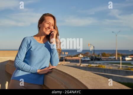 Vista laterale di giovane sognante afroamericana barefooted femmina in outfit casual seduto sulla sedia guardando attraverso la finestra mentre si ascolta alla musica in earp Foto Stock