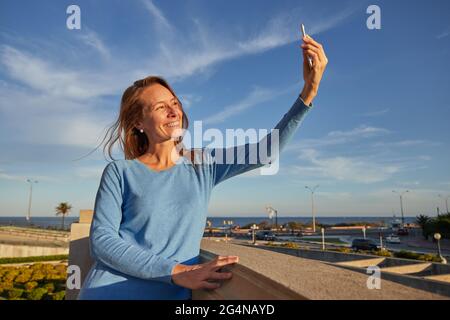 Donna adulta sorridente che prende selfie mentre si appoggia sulla recinzione vicino all'oceano in città strada in giornata di sole Foto Stock