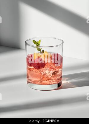 Bicchiere con cocktail a cubetti di ghiaccio e lamponi con sprig di menta posto su una superficie bianca Foto Stock
