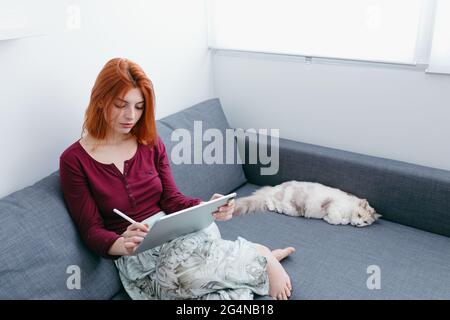 Giovane donna a piedi nudi con capelli rossi che naviga su internet su tablet mentre si siede con gatto sul divano a casa Foto Stock