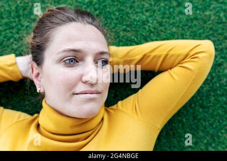 Dall'alto di sognante donna adulta in abiti casual giacente su erba verde con le mani dietro la testa e guardando via Foto Stock