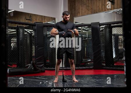 ritratto di uomo in nero sportswear preparazione per la lotta dura, fasciando pugno in sport bendaggi protettivi. in piedi su anello per prepararsi per mma figh Foto Stock