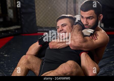 Due uomini boxer in guanti stanno combattendo in palestra usando grapping, seduto sul tappetino in anello in palestra. Concetto di kickboxing e sport Foto Stock