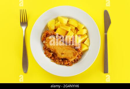 Patate al forno, carne di maiale e cavolo in piatto bianco su tavola gialla con posate, vista forma sopra Foto Stock