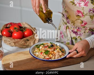 le mani della donna leccano l'olio d'oliva sul piatto di insalata di quinoa in cucina domestica, fondo di sacchetto di carta con pomodori e cassetti bianchi della cucina Foto Stock